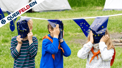 Drei Kinder halten sich EU-Flaggen vor das Gesicht, im Hintergrund ein Lager