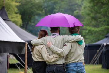 Zwei Pfadis und ein Leiter mit Kluft stehen Arm in Arm mit dem Rücken zum Bild und halten einen aufgespannten Regenschirm über sich. Im Hintergrund ist ein Lager mit Schwarzzelten zu sehen. 