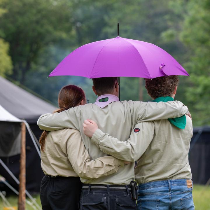 Zwei Pfadis und ein Leiter mit Kluft stehen Arm in Arm mit dem Rücken zum Bild und halten einen aufgespannten Regenschirm über sich. Im Hintergrund ist ein Lager mit Schwarzzelten zu sehen. 